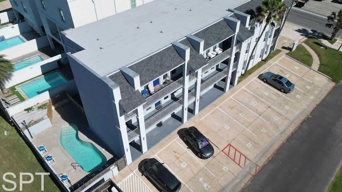 Un edificio de varios pisos con piscina en la azotea, estacionamiento con varios autos y un pequeño espacio con césped y una palmera.