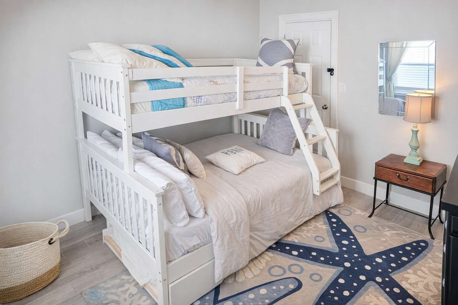 Una cama litera con un tapete estampado con motivos de estrellas de mar, una mesita de noche con lámpara y una cesta de mimbre.