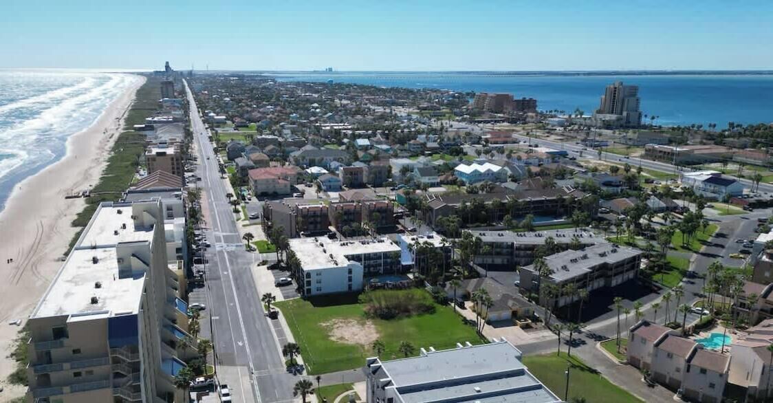 An aerial view of a coastal town with a beachfront, residential buildings, and a large hotel complex.