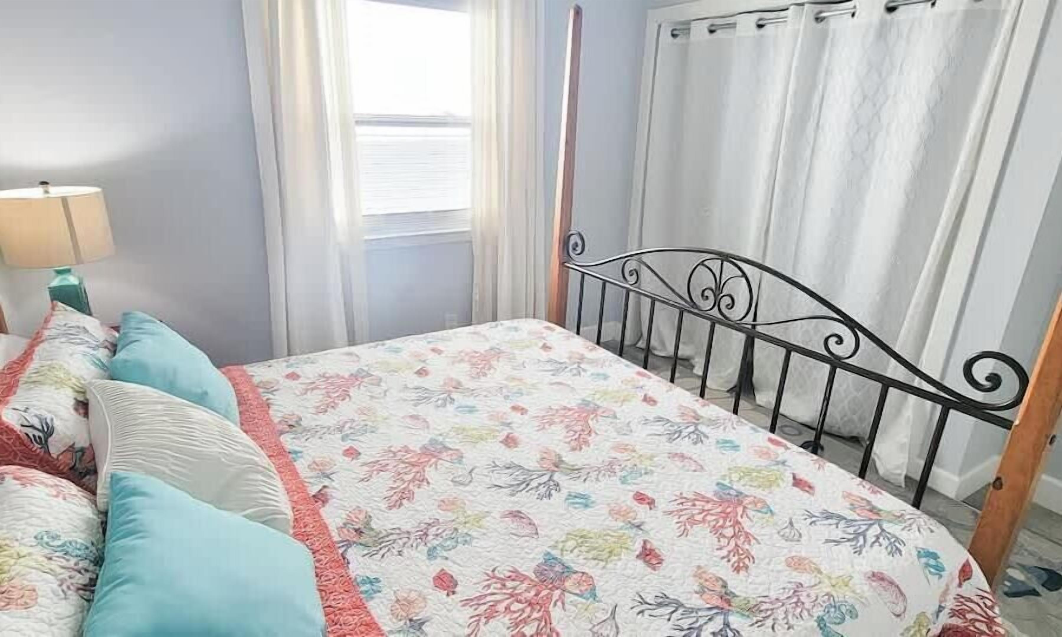 Une chambre à coucher avec un lit, des oreillers, une table de chevet, une lampe, une fenêtre avec des rideaux et une tête de lit en métal.