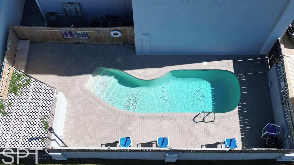 A swimming pool with a wavy edge, surrounded by a paved deck. There are lounge chairs and a small poolside table.