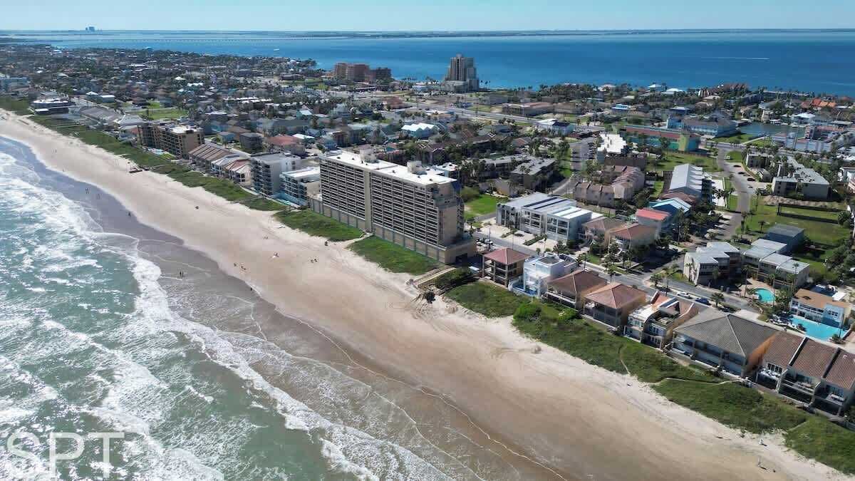 Eine Luftaufnahme einer Küstenregion mit Strand, Wohnhäusern und einem Hotel mit mehreren Etagen und Balkonen.