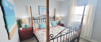 A bedroom with a four-poster bed, two bedside tables with lamps, a wooden headboard, a wall-mounted painting, and a window with curtains.