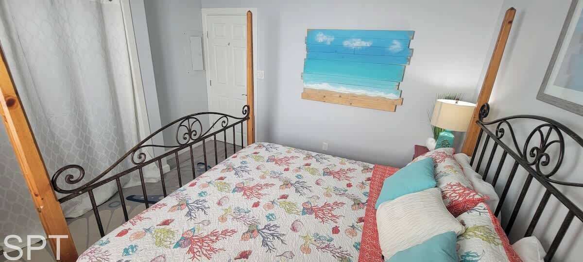 A room with a bed, a window with curtains, a wall-mounted shelf with a painting, and a bedside table with a lamp.