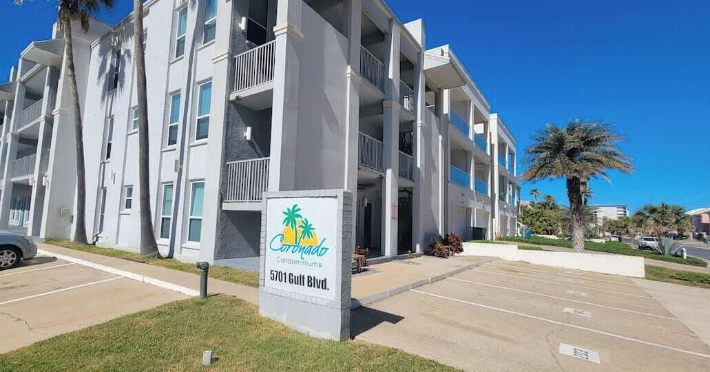 A multi-story building with balconies, a sign for Coronado Condominiums at 5701 Gulf Blvd., and a parking lot.