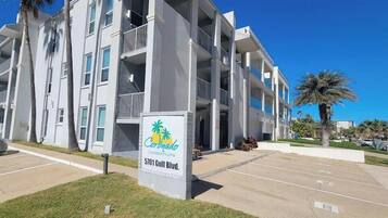 A multi-story building with balconies, a sign for Coronado Condominiums at 5701 Gulf Blvd., and a parking lot.