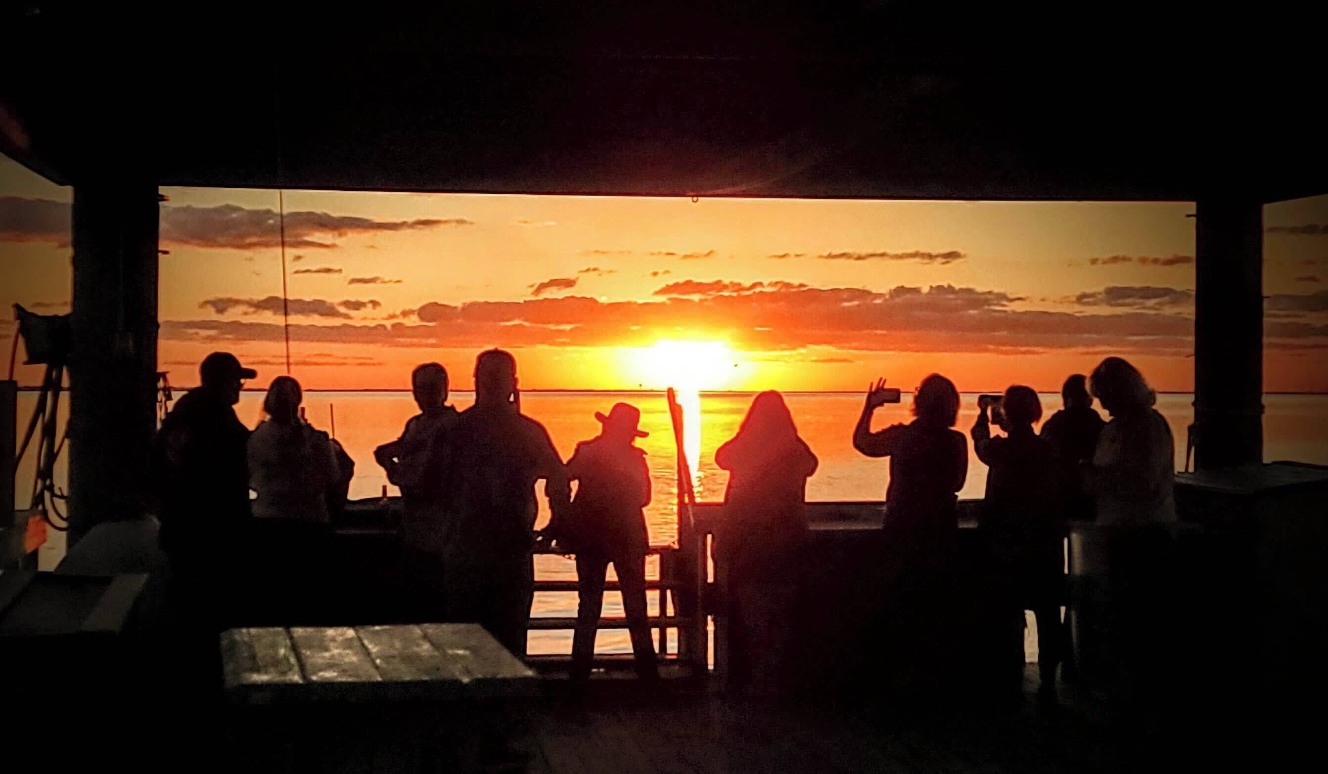 Um grupo de pessoas silhuetadas contra o pôr do sol em um píer, com vista para o oceano.