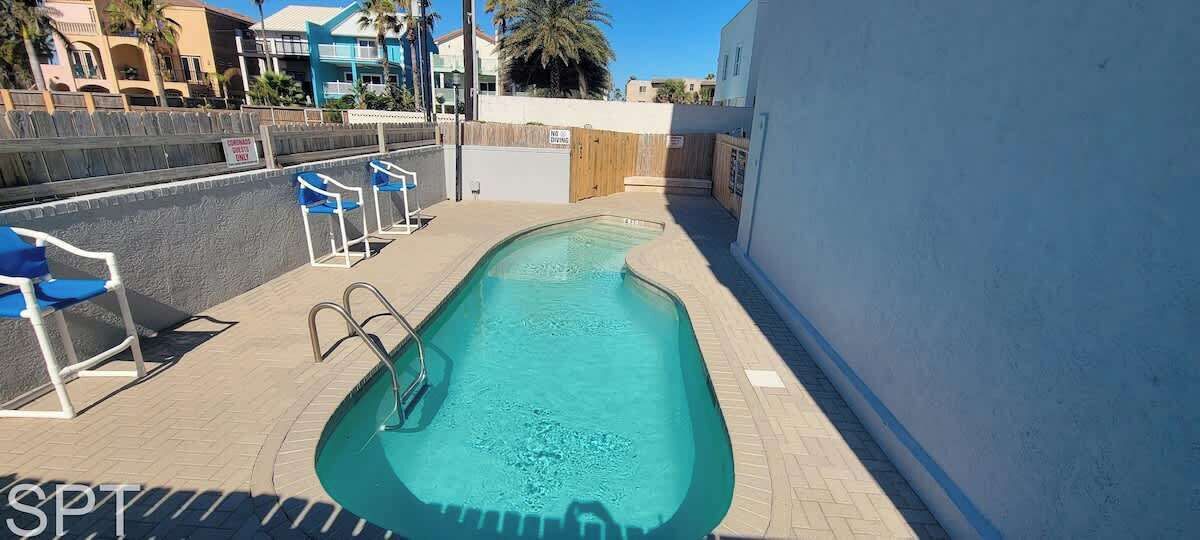 A swimming pool with a diving board, surrounded by a low brick wall and blue chairs.
