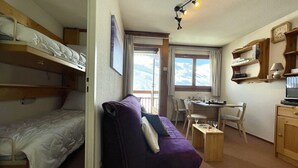 A compact room with bunk beds, a purple sofa, a small table, and a desk with chairs. - (7.B17) Appartement skis aux pieds Les Menuires (Les Menuires)