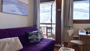 A room with a purple sofa, a dining table with chairs, and a window with a snowy view. - (7.B17) Appartement skis aux pieds Les Menuires (Les Menuires)