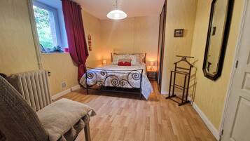 Une chambre à coucher comprenant un lit, une chaise, un radiateur, une fenêtre avec des rideaux, une table de chevet avec une lampe et un miroir accroché au mur.