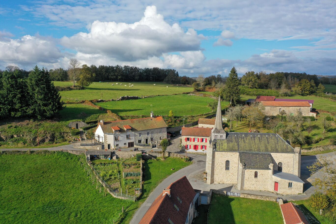 Een landelijk dorp met stenen gebouwen, waaronder een kerk met een hoge spits, omringd door groene velden en bomen.