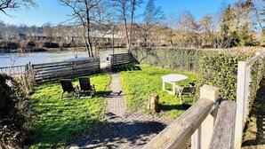 En trädgård vid floden med ett trädäck, stolar och ett bord.
