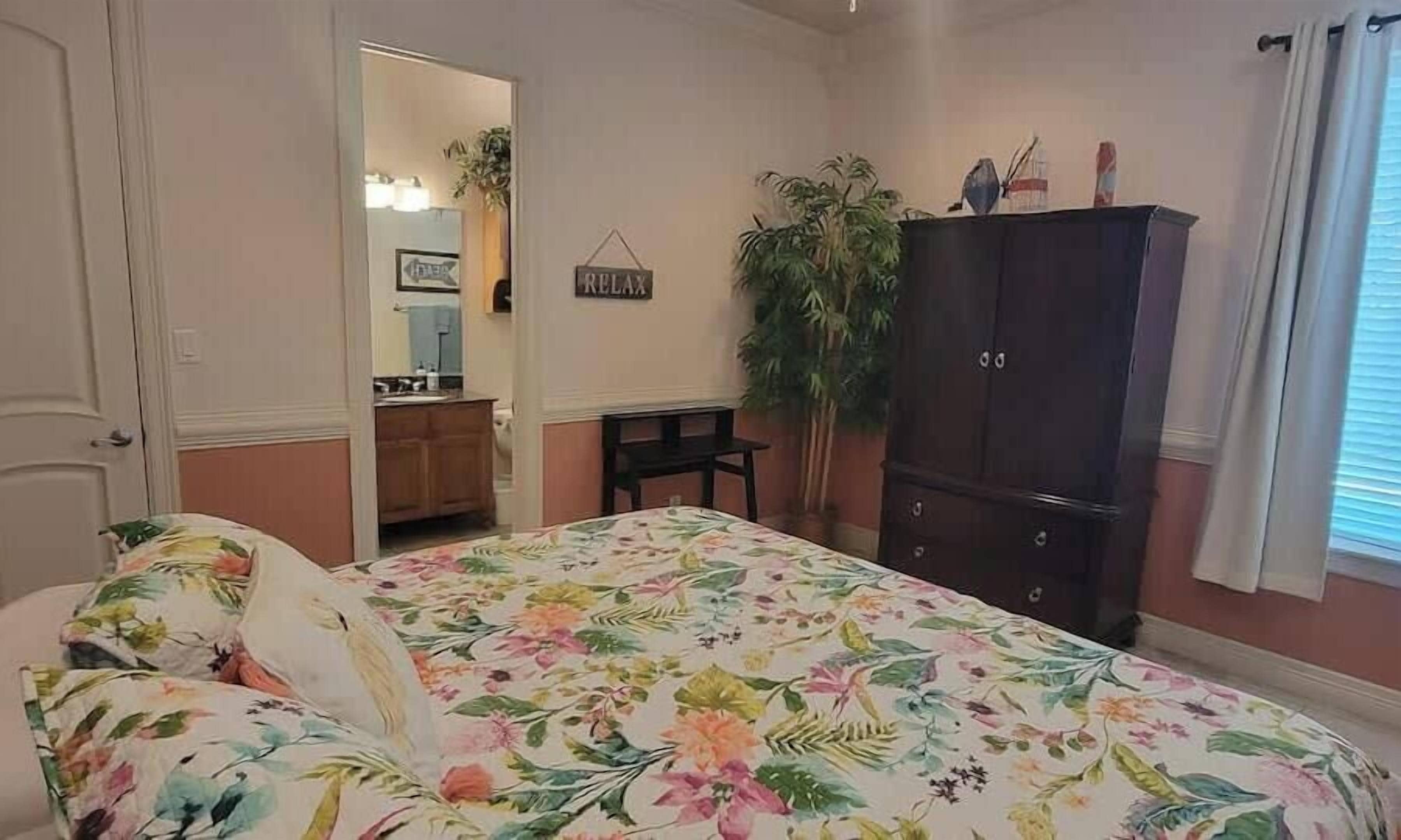 A bedroom with a floral bedspread, a dark wooden wardrobe, a small table, and a potted plant.