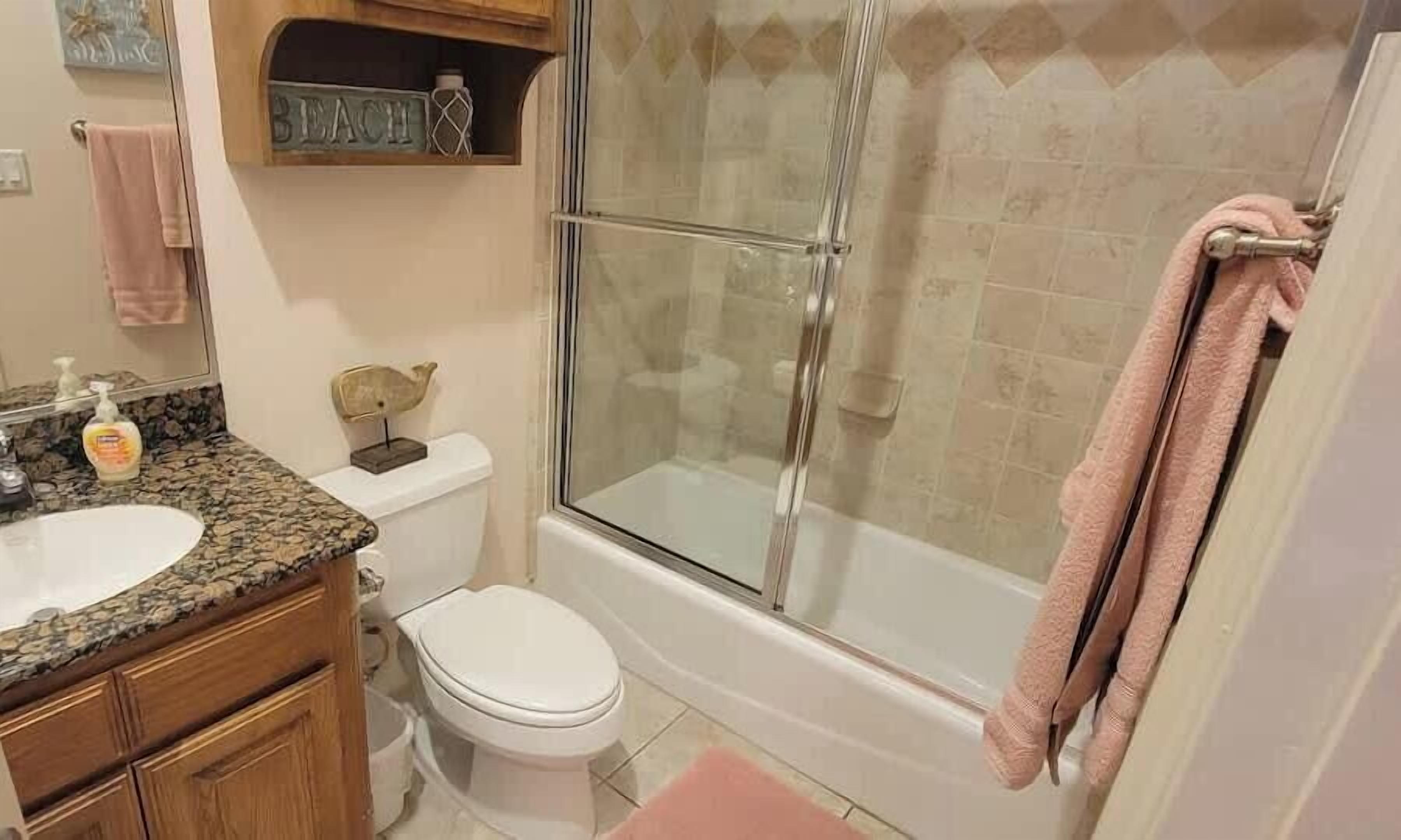 Un baño con lavamanos de encimera de granito, inodoro, ducha con mampara de cristal y una toalla rosa colgada en la pared.