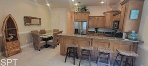 A kitchen with wooden cabinets, a central island with stools, a dining area with wicker chairs, and a decorative wooden piece on the wall. - Island Time #4 (South Padre Island)