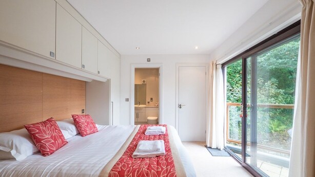 Une chambre d’hôtel moderne avec un grand lit, une tête de lit en bois, une salle de bain visible à travers une porte ouverte et une grande fenêtre avec des rideaux.