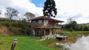 Una casa de dos pisos con balcón, un estanque al frente y una zona arbolada.