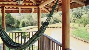 A wooden deck with a hammock, wooden railing, and a view of a river and greenery. - Chalé sol nascente  (Campo Alegre)