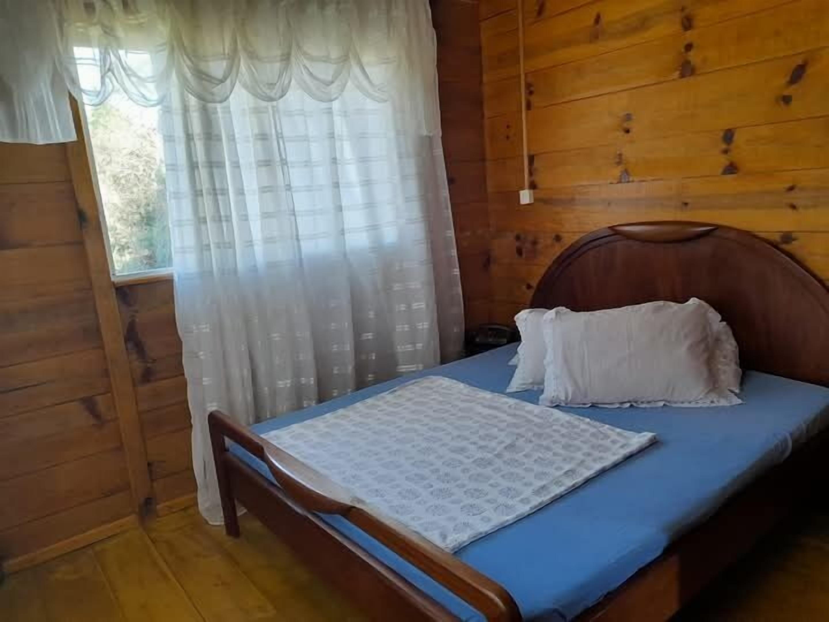 Habitació de cabana de fusta amb un llit, una finestra amb cortines lleugeres i un panell de fusta al capçal.