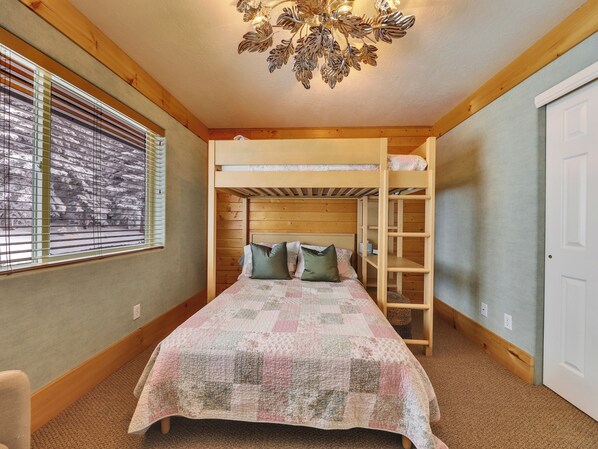 A bunk bed room with a wooden ceiling, a quilted bedspread, and a window with blinds.