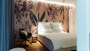 A bedroom with a bed, a chair, a nightstand, and a wall with a floral pattern. - The Kepos Hotel Erlangen (Erlangen)
