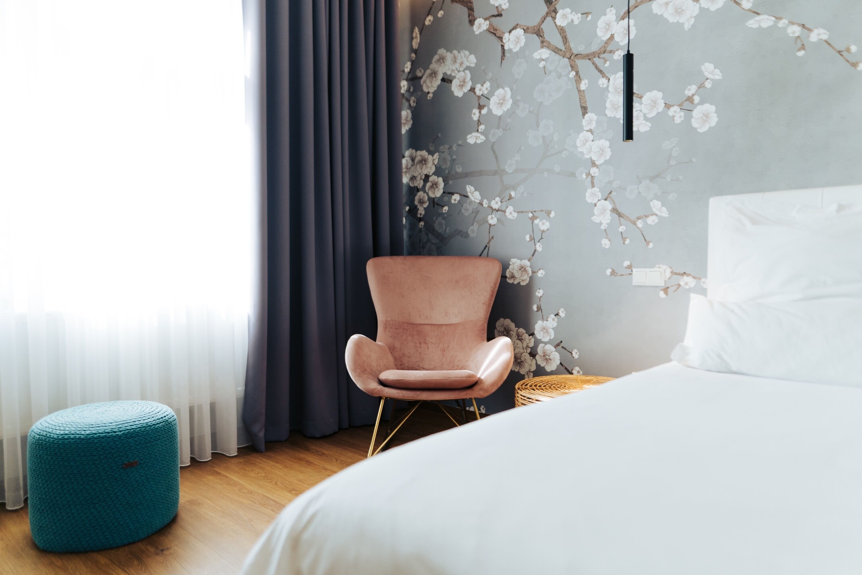 A bedroom with a bed, a pink chair, a teal ottoman, and a wall with floral wallpaper.
