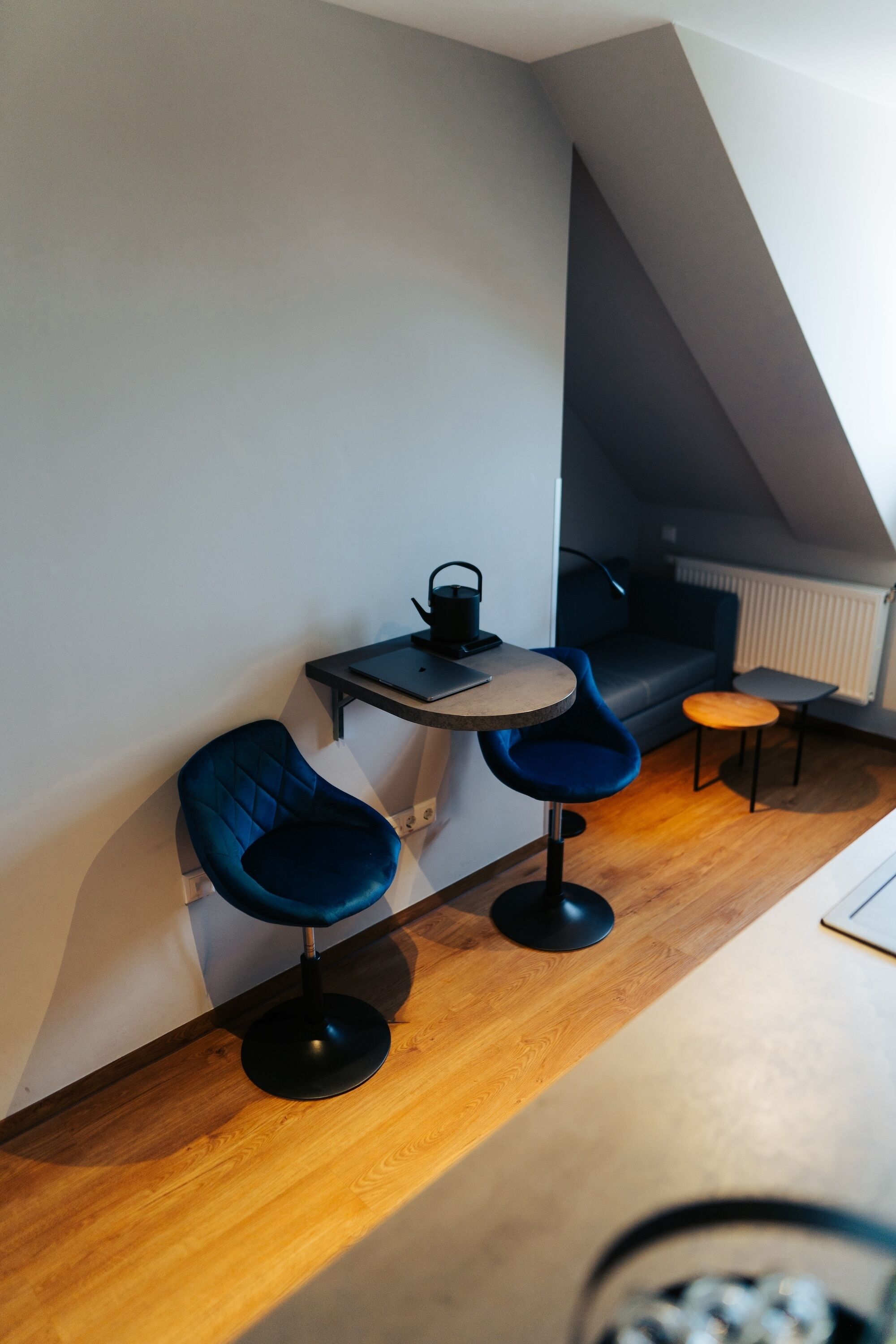 A modern room with a loft-style ceiling, a small table with two blue chairs, a sofa, and a radiator.