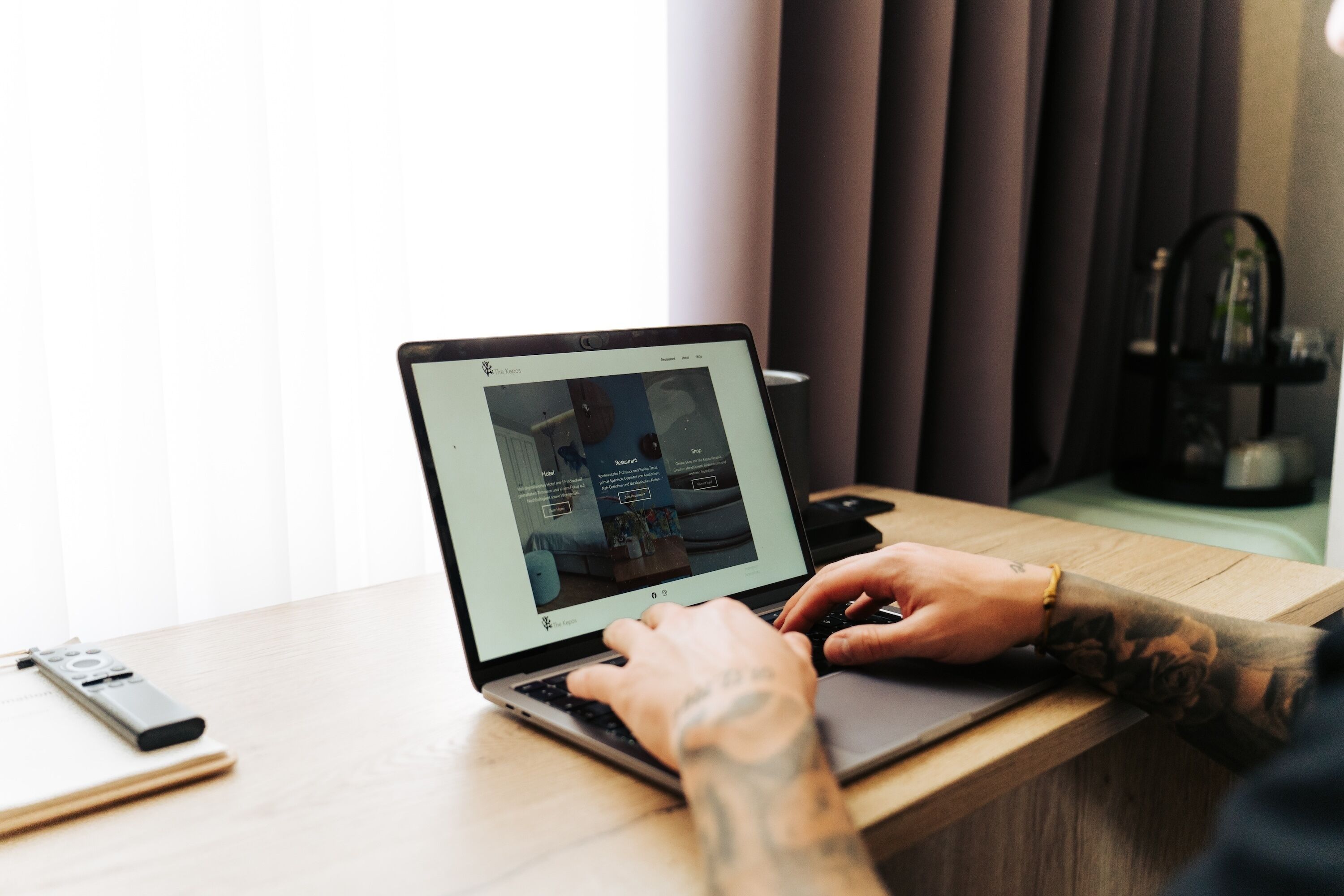 A person using a laptop on a wooden table with a remote control and a smartphone beside it.