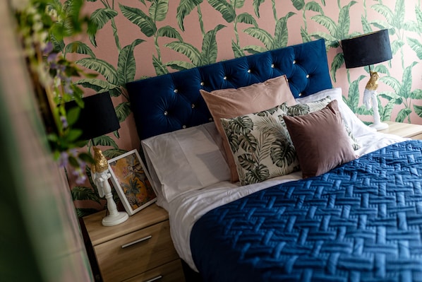 A bedroom with a blue bedspread, a tufted headboard, and a wall with a leafy pattern.
