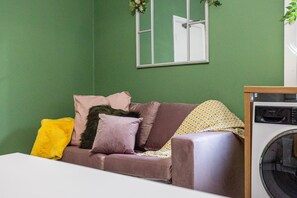 A room with a green wall, a brown sofa with cushions, a yellow throw, and a washing machine.