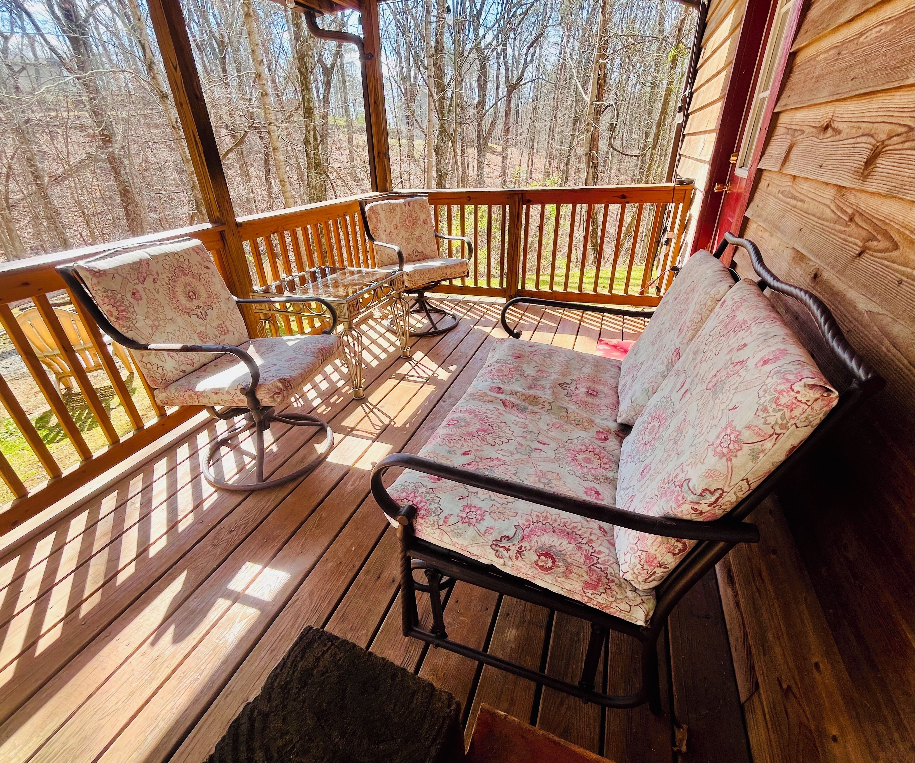 A wooden deck with a sofa and chairs, a glass table, and a view of trees.