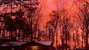 Une cabane dotée d’une véranda et de deux chaises, entourée d’arbres, avec un ciel rouge en arrière-plan.
