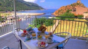 A breakfast table with various food items and drinks on a balcony overlooking a coastal area with a mountain and a beach.
