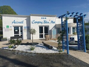 A building with "Accueil" and "Camping Bleu Noir" signs, a bench, and a blue pergola structure.