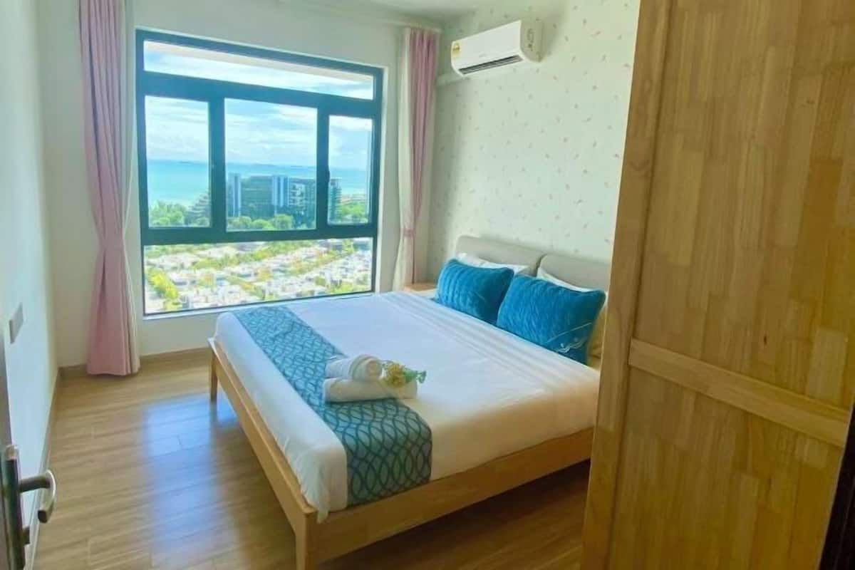 A hotel room with a large bed, a view of a cityscape through a window, and a wooden wardrobe.