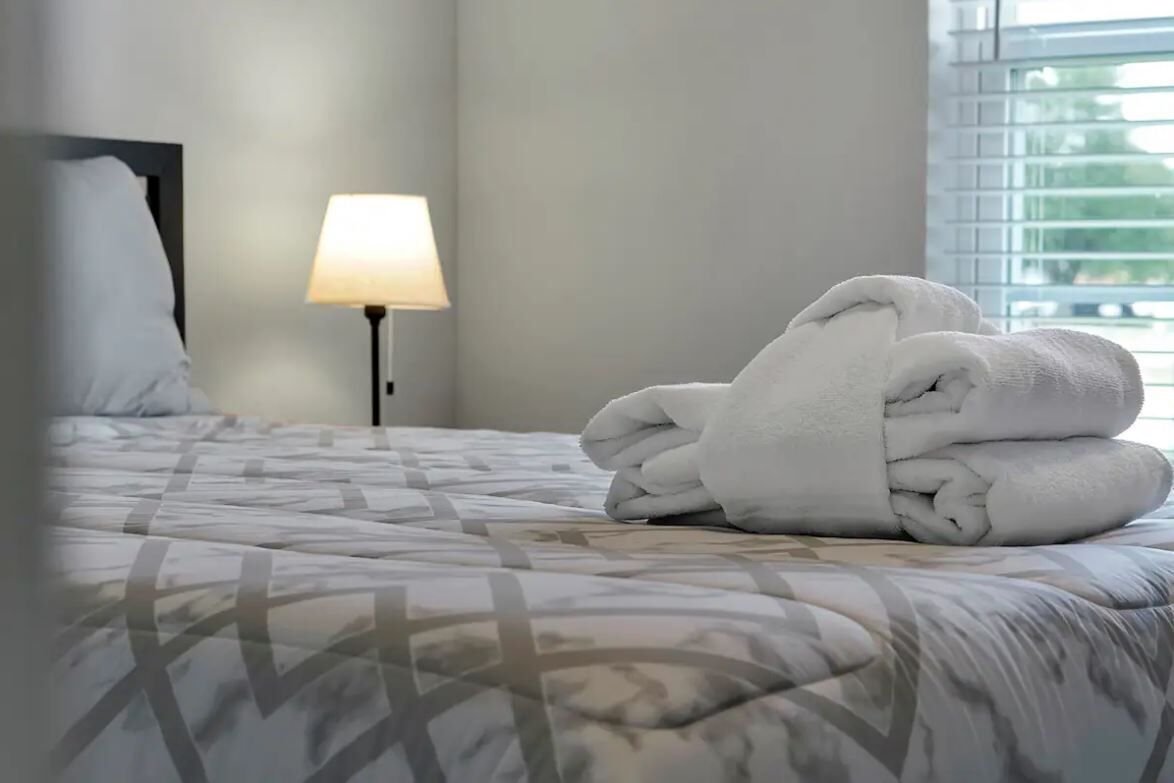 A neatly made bed with a patterned bedspread, a stack of folded towels, a bedside lamp, and a window with blinds.