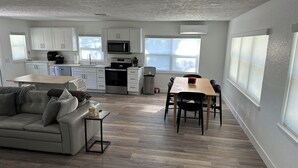 Una cocina moderna con armarios blancos, microondas, horno y cafetera. Hay una zona de comedor con una mesa y sillas de madera, y un sofá gris con cojines.