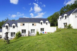 Un bâtiment blanc avec un toit en pente, une terrasse avec des meubles d’extérieur et une pelouse devant.