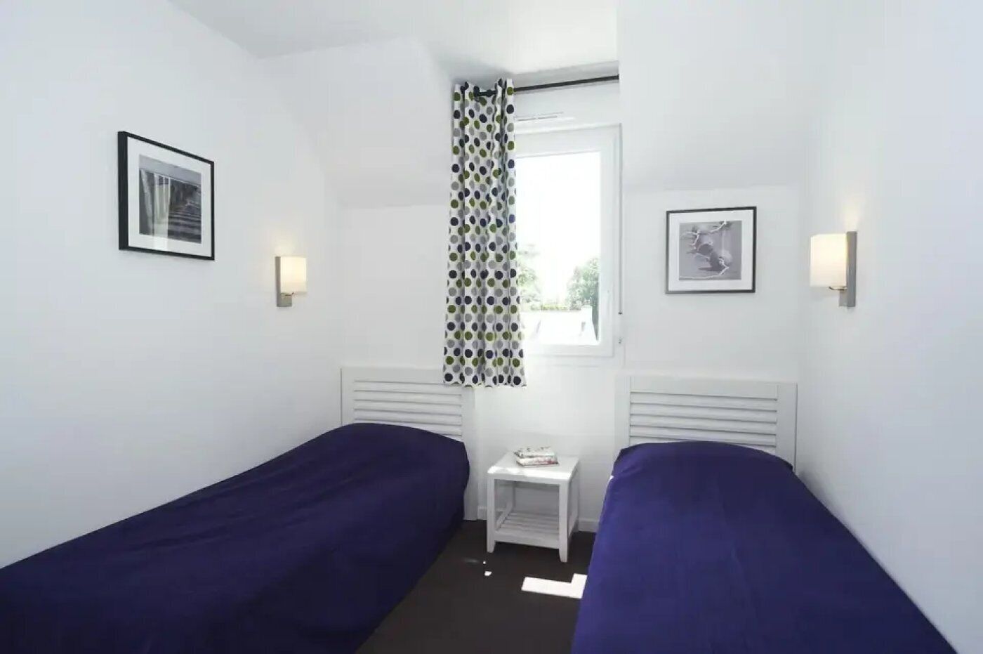 Une chambre avec deux lits simples, une petite table, une fenêtre avec un rideau et deux lampes fixées au mur.