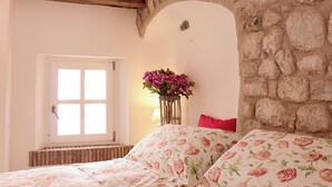 A bedroom with a stone wall, a window, a bed with floral bedding, and a vase of flowers. - Cà BIANCA Residence - REFOSCO Apartment - Historical Center POLCENIGO (Polcenigo)