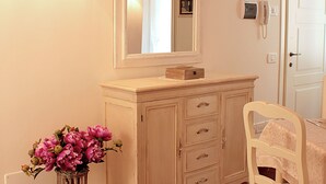 A room with a wooden floor, a white dresser with drawers, a mirror, a vase with flowers, a table with a chair, and a wall-mounted telephone. - Cà BIANCA Residence - REFOSCO Apartment - Historical Center POLCENIGO (Polcenigo)