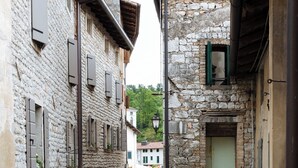 A narrow cobblestone street flanked by stone buildings with shutters, a metal grate on the ground, and a view of a distant building. - Cà BIANCA Residence - REFOSCO Apartment - Historical Center POLCENIGO (Polcenigo)