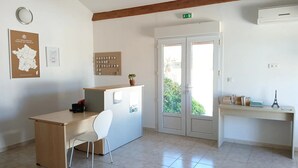Een kamer met een bureau, stoel en een kleine plant in een pot. Er is een deur met ruitpanelen, een luchtverkoeler aan de muur en een kaart.