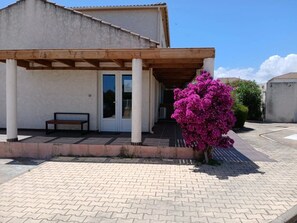 Un bâtiment doté d’un porche couvert, d’un banc et d’un arbuste à fleurs roses.