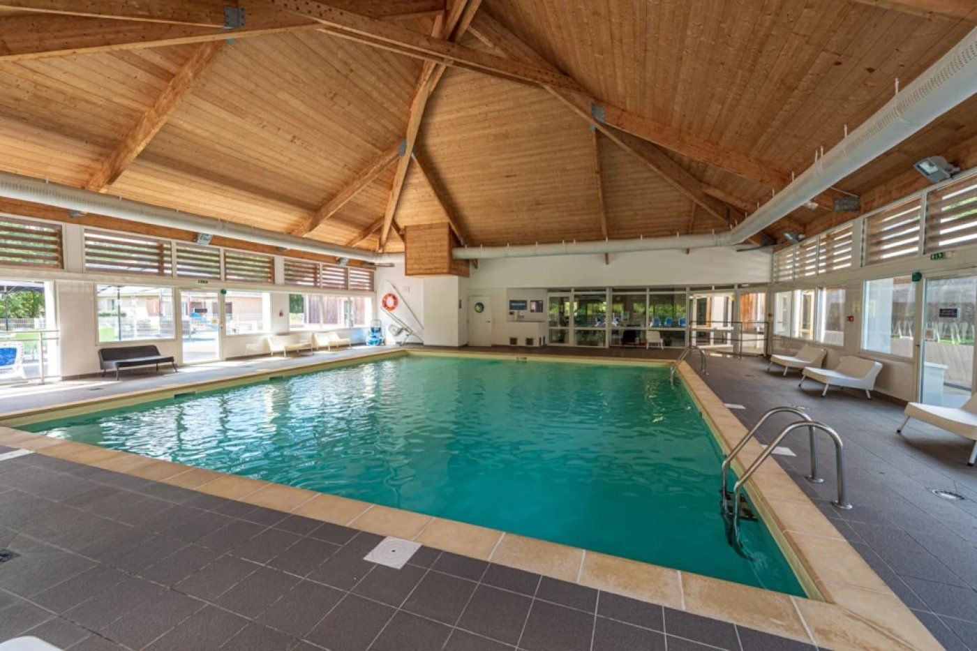 An indoor swimming pool with a wooden ceiling, tiled floor, and seating areas.