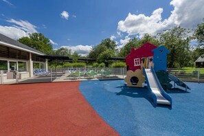 Un parque infantil con zona de juegos roja y azul, un tobogán y un edificio con techo rojo.