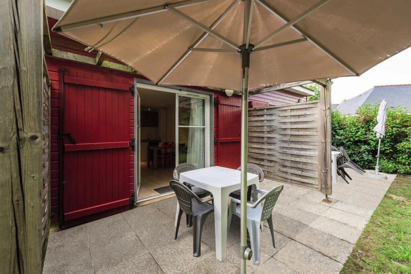 An outdoor patio with a table and chairs under a large umbrella, a red door, and a wooden fence in the background.