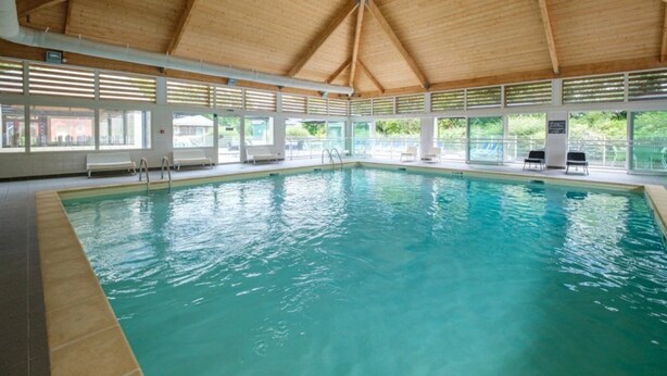 Una piscina interior con techo de madera, ventanas amplias y sillones de descanso.