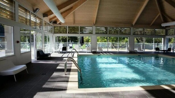 An indoor swimming pool with clear water, a wooden ceiling, and a view of the outdoors through large windows.
